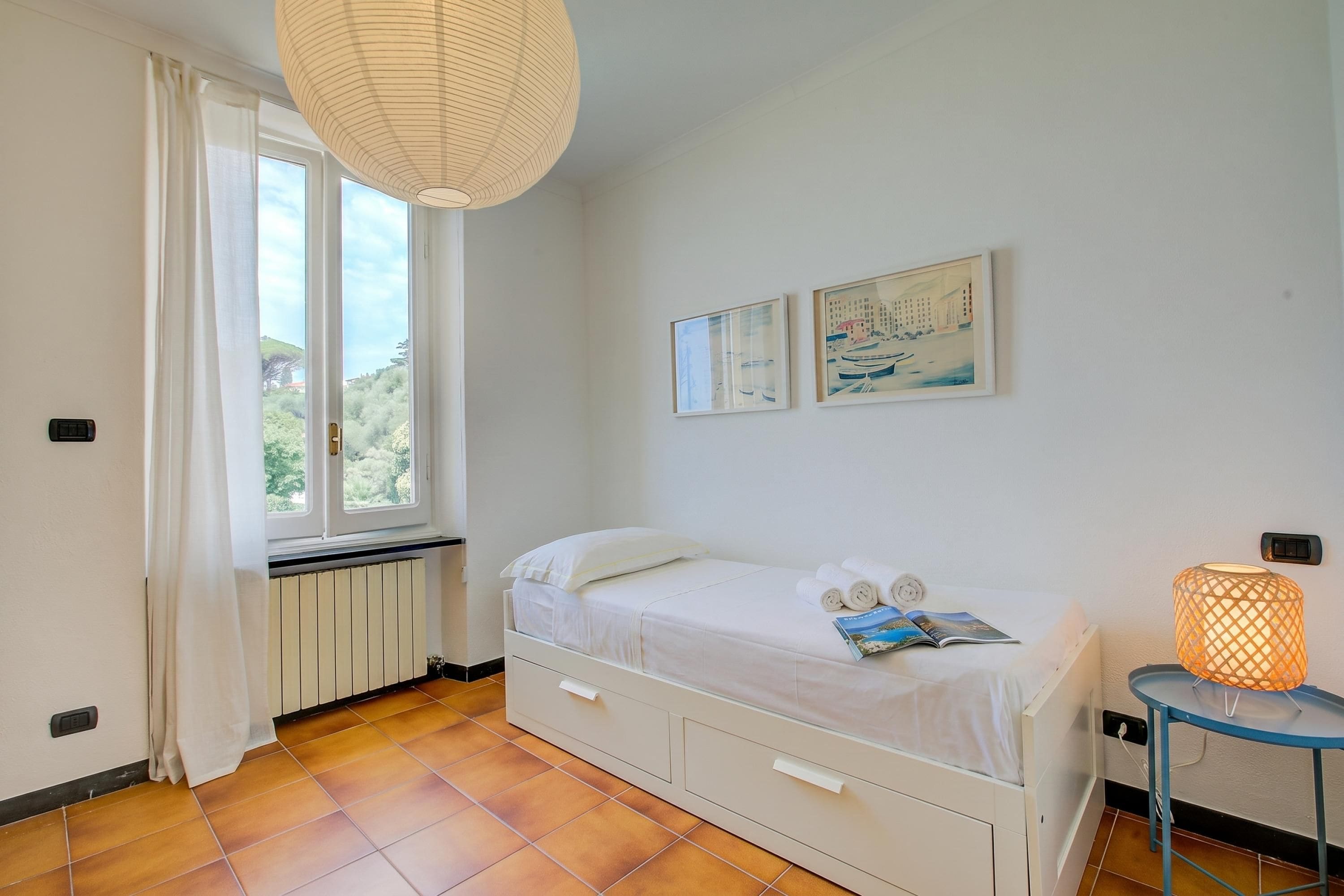 Une chambre à coucher comprenant un lit, un radiateur, une fenêtre avec des rideaux, une lampe fixée au mur et deux tableaux encadrés.