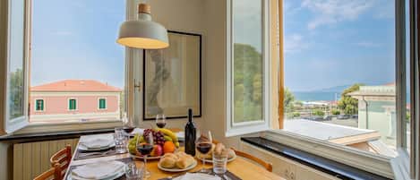 A dining table set with plates, glasses, and a fruit bowl, overlooking a coastal view.