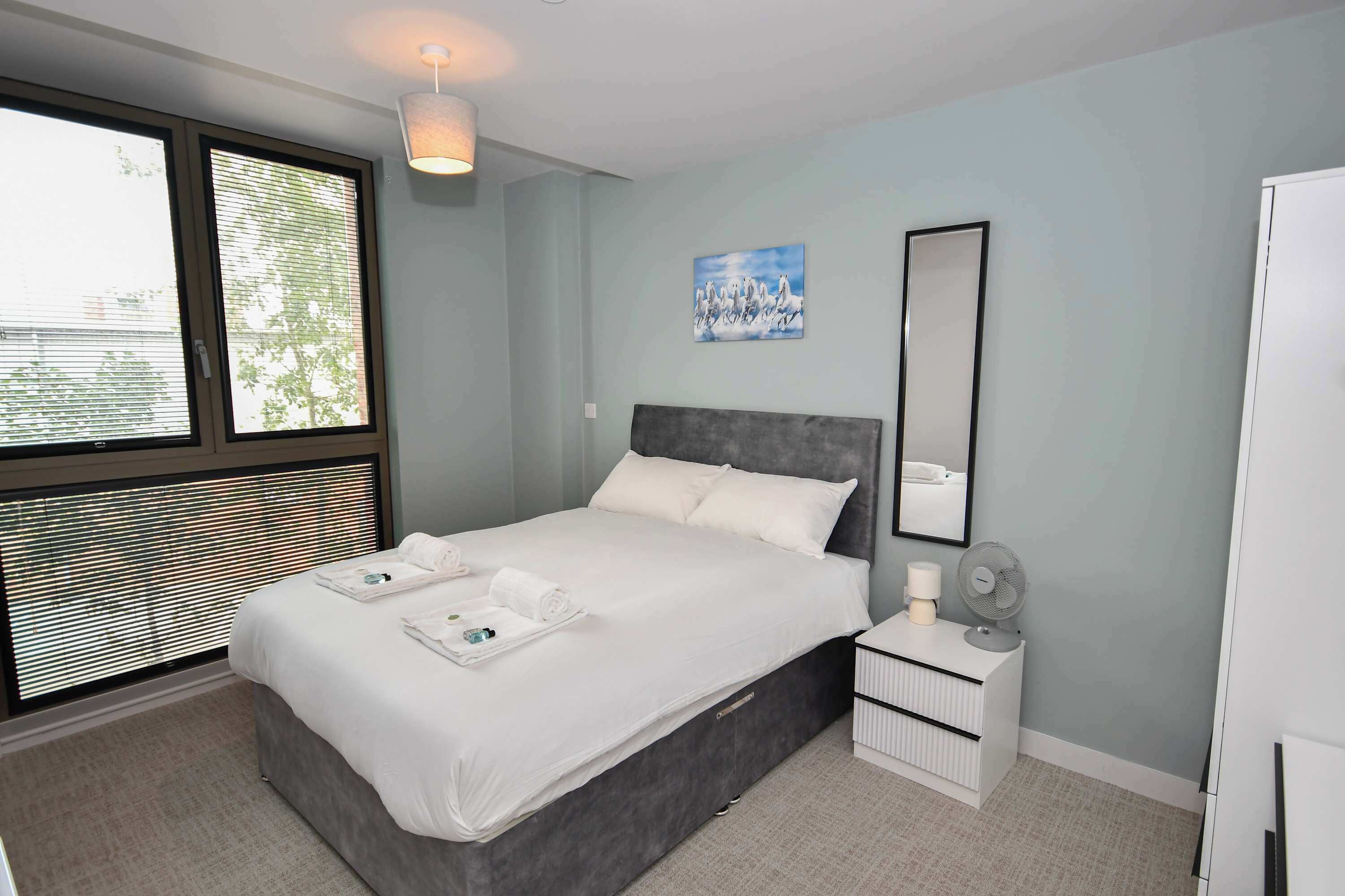 Una habitación de hotel moderna con una cama grande, una mesita de noche con lámpara, un espejo y una ventana con persianas.