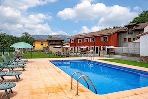 Ein Swimmingpool mit Liegestühlen und Sonnenschirmen, umgeben von Gebäuden und einer Bergkulisse.