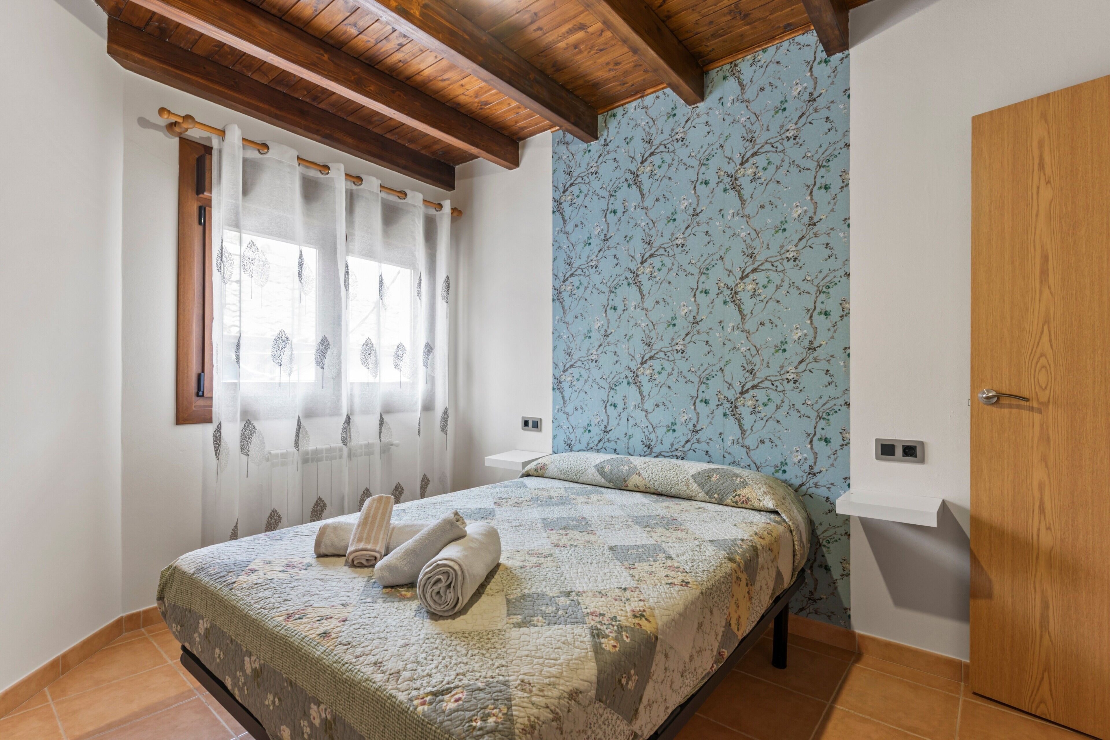 A bedroom with a bed, curtains, a wooden ceiling, a floral wallpaper, and a wooden wardrobe.