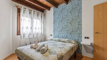 A bedroom with a bed, curtains, a wooden ceiling, a floral wallpaper, and a wooden wardrobe.