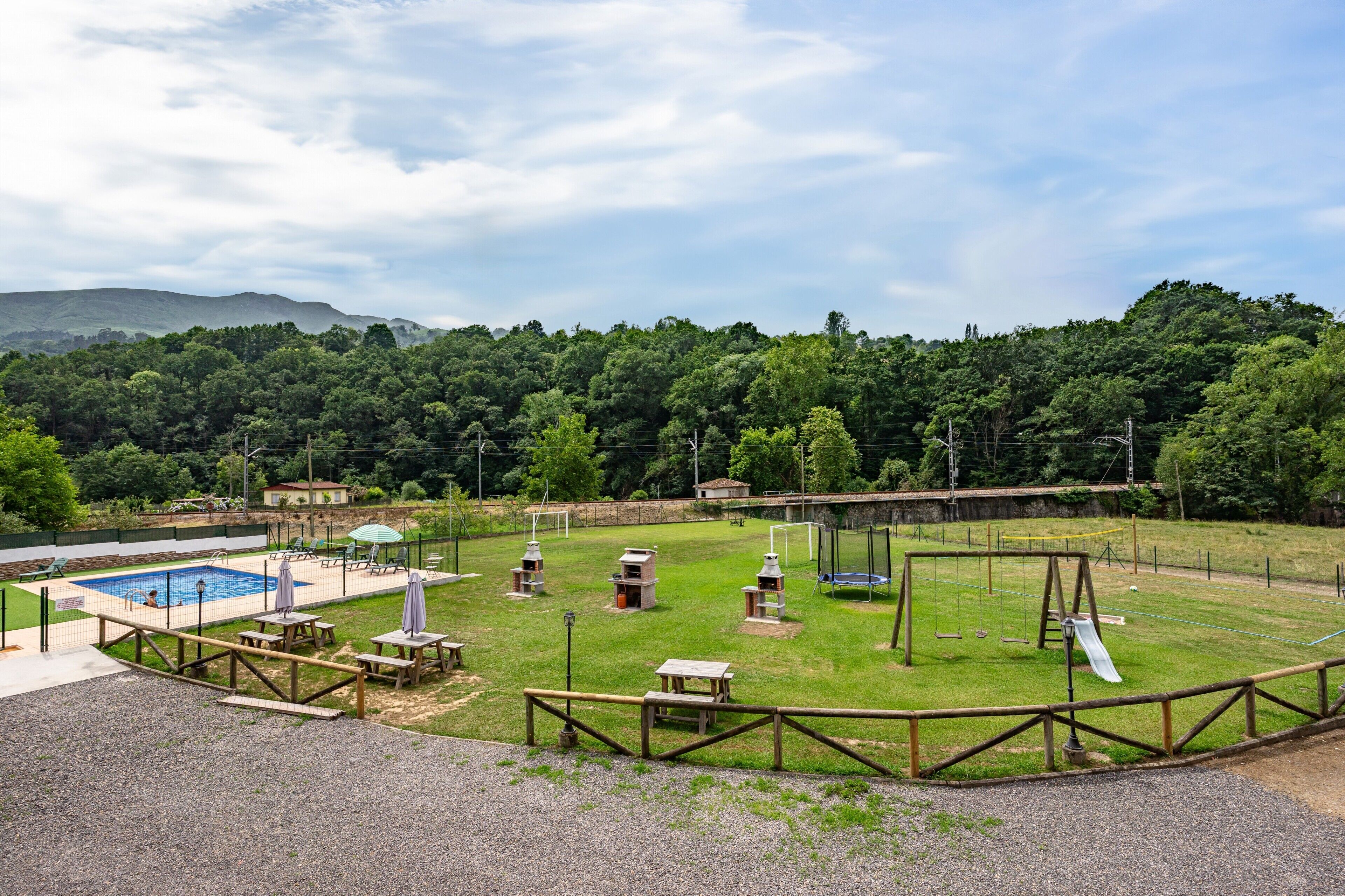 A fenced outdoor area with a pool, picnic tables, a swing set, and a playground.