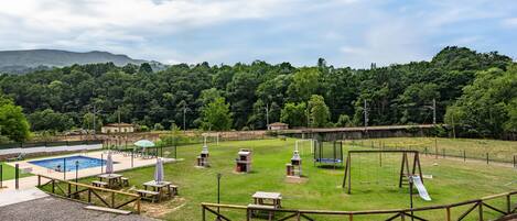 A fenced outdoor area with a pool, picnic tables, a swing set, and a playground.