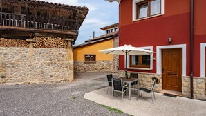 A courtyard with a stone wall, a wooden fence, a red building with a door and window, and a table with chairs under an umbrella.