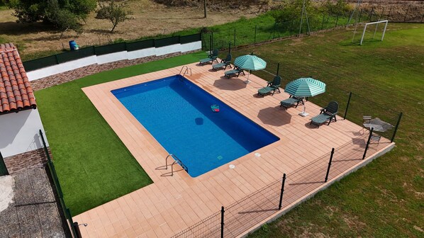 A rectangular swimming pool with a tiled deck, surrounded by a fence, and equipped with lounge chairs and umbrellas.