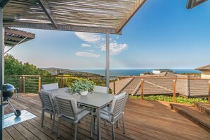 A covered outdoor deck with a table and chairs, overlooking a scenic view of the ocean and hills. - Pezula Sunset Views MG10 (Knysna)