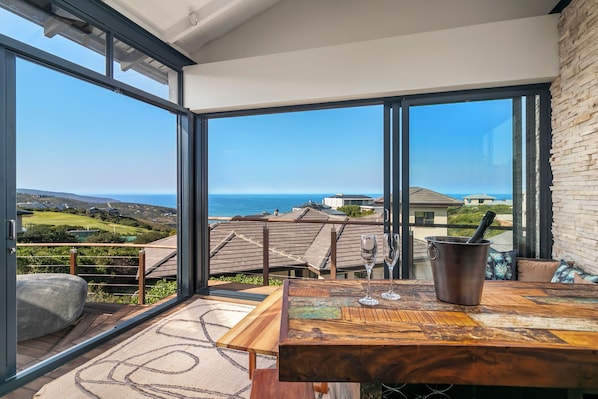 A room with large windows, a wooden table, a glass, and a metal container. - Pezula Sunset Views MG10 (Knysna)