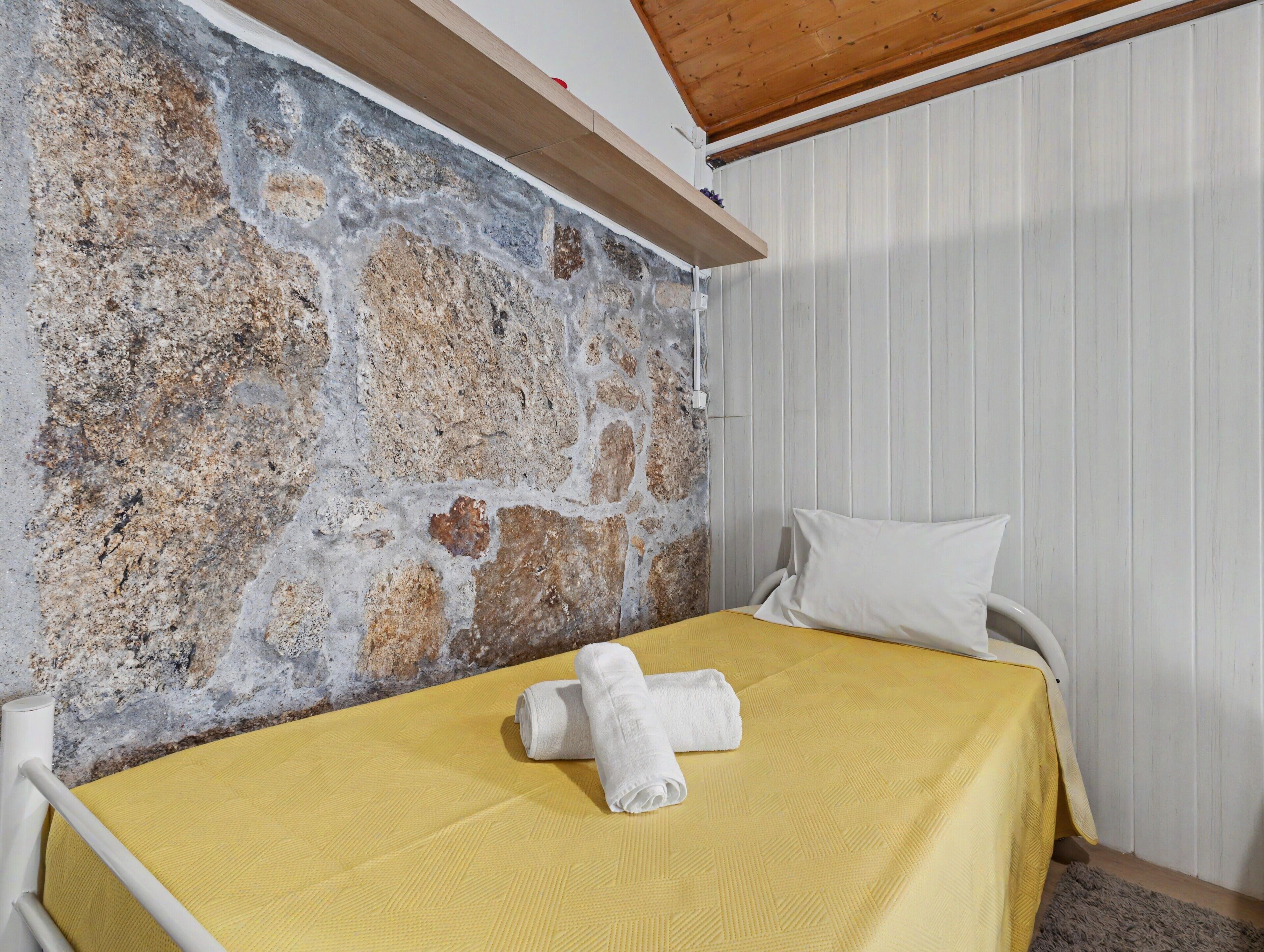 A room with a stone wall, a wooden ceiling beam, a bed with a yellow bedspread, and a white pillow.