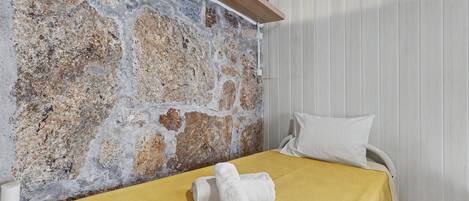 A room with a stone wall, a wooden ceiling beam, a bed with a yellow bedspread, and a white pillow.