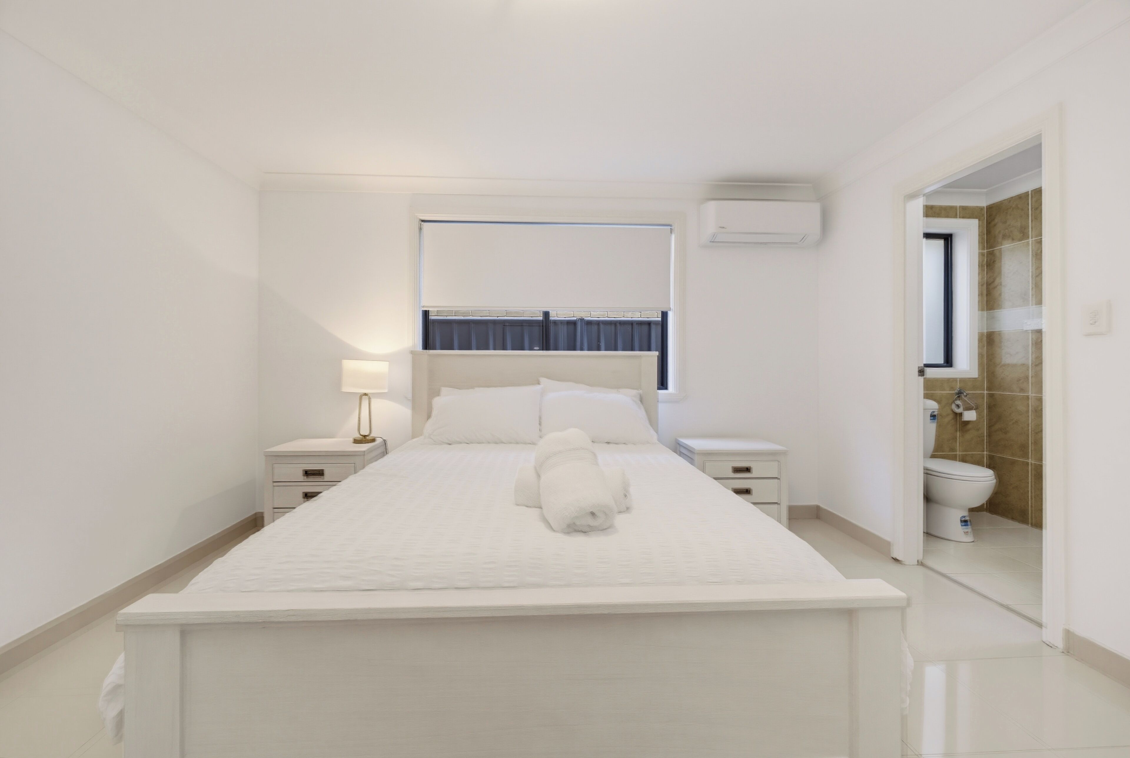 A hotel room with a bed, bedside tables, a lamp, a window, and a bathroom visible through an open door.