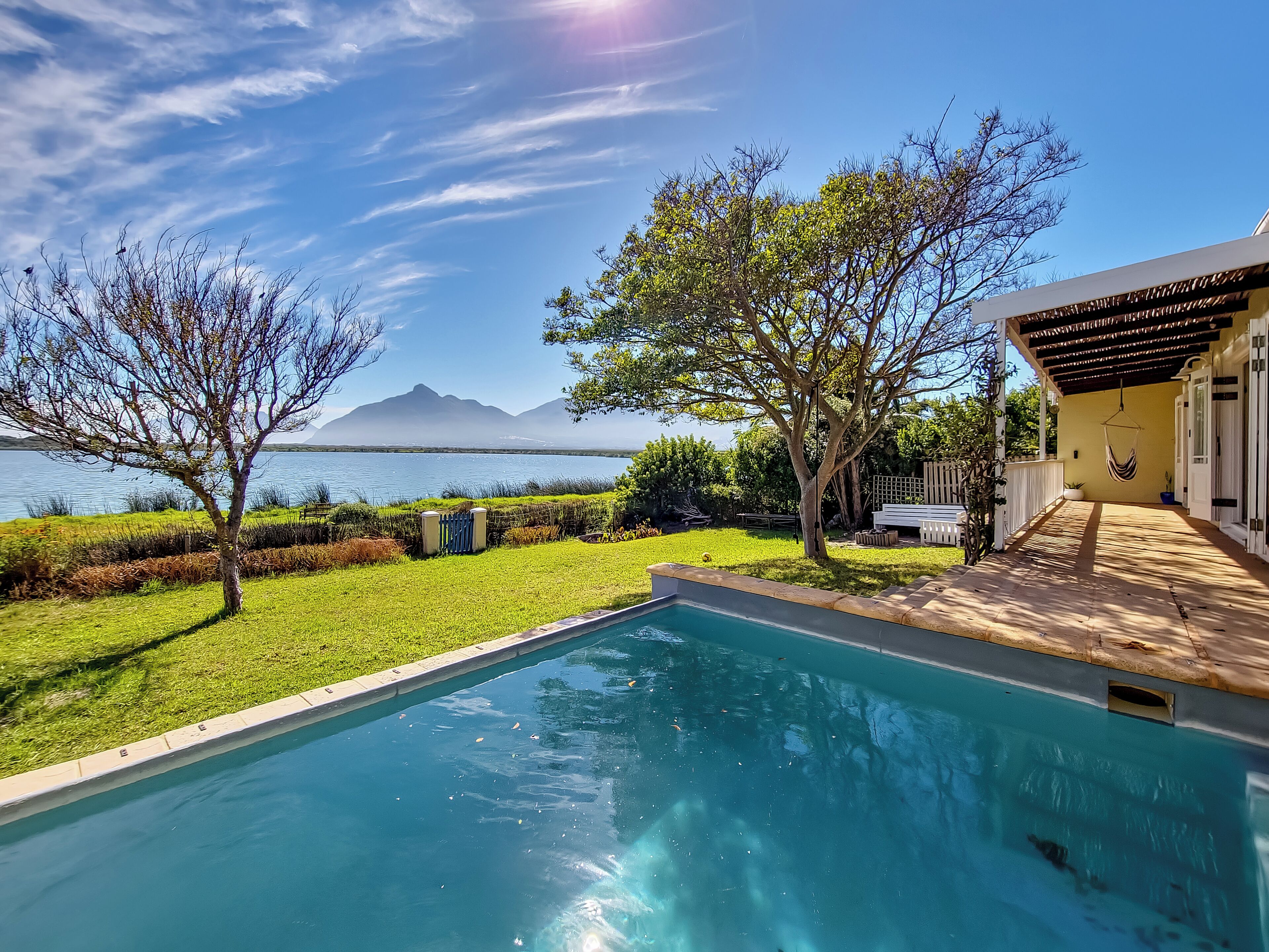 A swimming pool with a view of a lake and mountains, surrounded by a well-maintained lawn and a house with a covered patio.