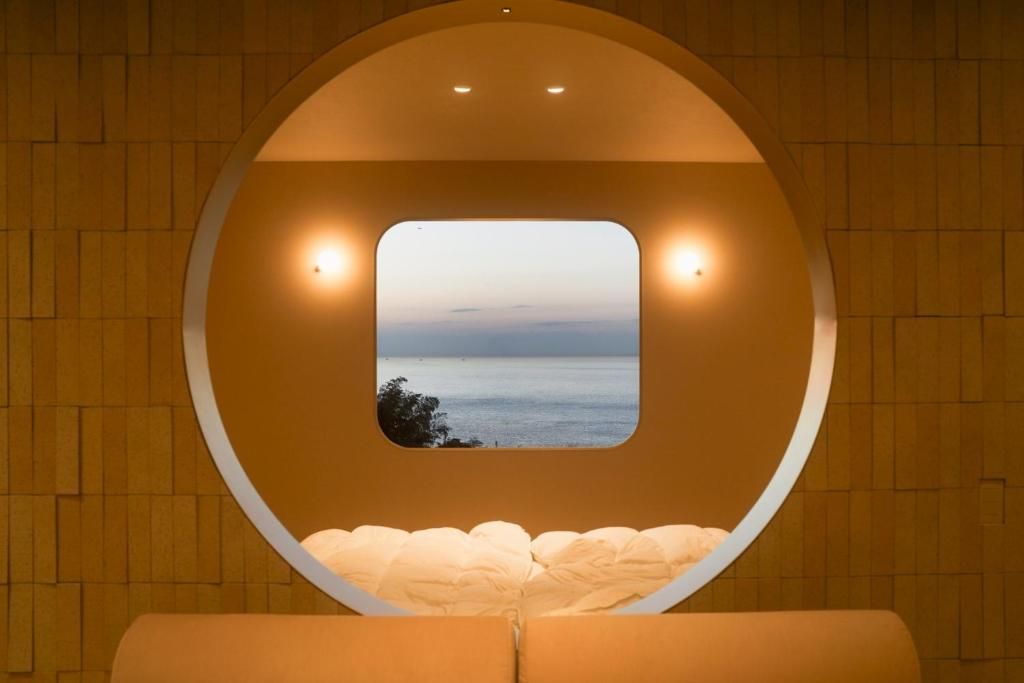 A circular room with a view of the sea through a window, a bed, and two wall-mounted lights.