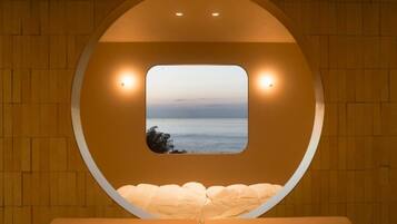 A circular room with a view of the sea through a window, a bed, and two wall-mounted lights.