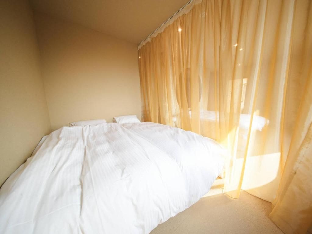 Una cama con ropa blanca en una habitación con paredes color beige y cortinas traslúcidas.