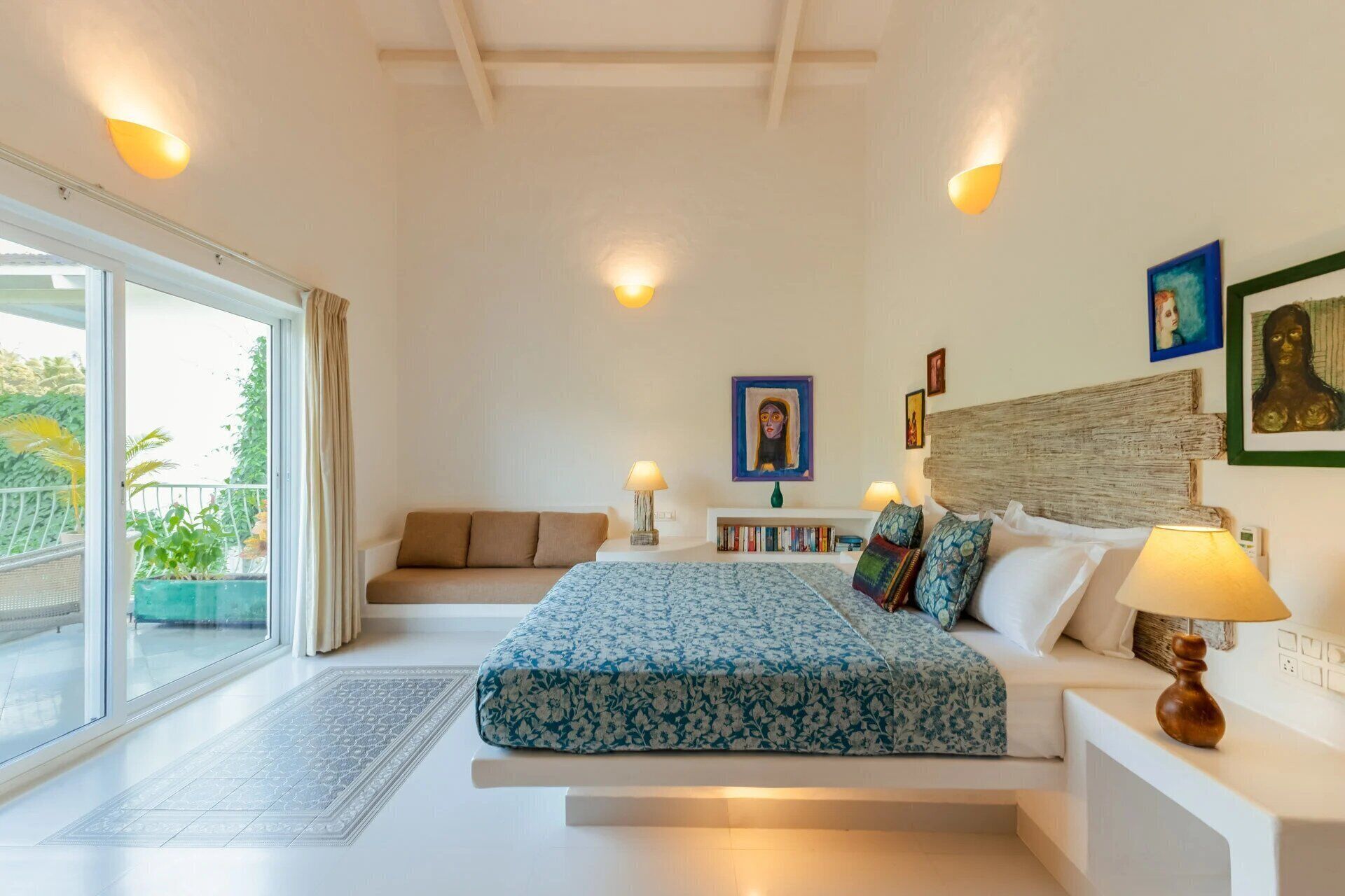 A bedroom with a bed, bedside tables, a sofa, a window with curtains, and artwork on the walls.