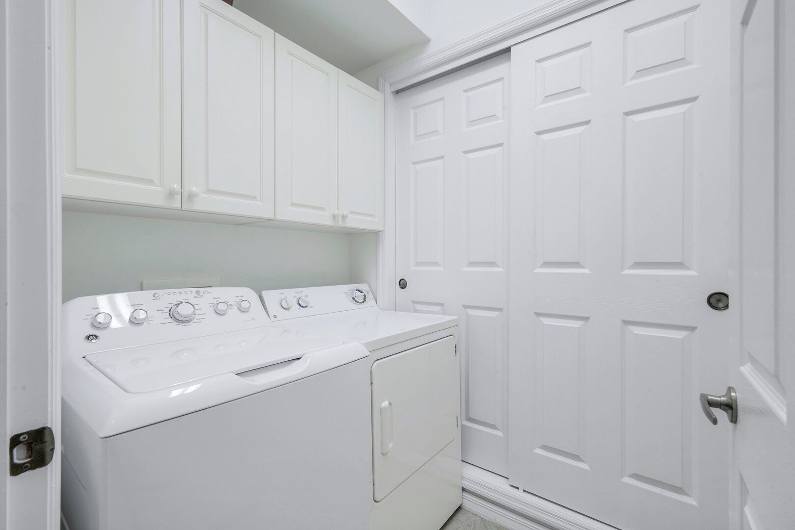 A laundry room with a front-loading washer and dryer, white cabinets above, and a white door with a silver handle.
