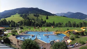 A swimming pool with a water slide, surrounded by green hills and clear skies. - Cozy Holiday Apartment With Balcony Near ski Slope (Maria Alm am Steinernen Meer)