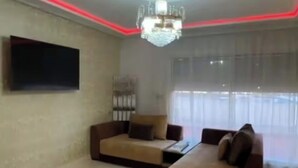 Een woonkamer met een kroonluchter, banken, een flatscreen televisie en een groot raam met gordijnen.