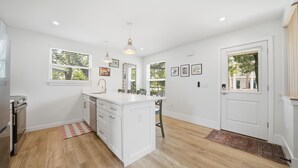 A modern kitchen with white cabinetry, a central island, and a dining area with a table and chairs. - Pelican's Perch Cottage - New & Modern In Perfect Location w/ Waterfront Deck (St. Augustine)