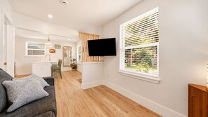 A modern living room with a grey sofa, a flat-screen TV mounted on the wall, and a large window with blinds. - Pelican's Perch Cottage - New & Modern In Perfect Location w/ Waterfront Deck (St. Augustine)
