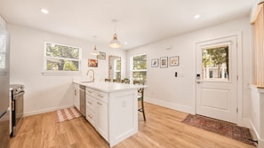 A modern kitchen with white cabinetry, a central island, and a dining area with chairs. - Pelican's Perch Cottage - New & Modern In Perfect Location w/ Waterfront Deck (St. Augustine)