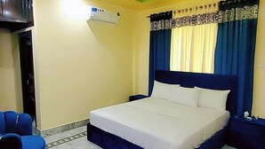 Habitación de hotel con cama, ventilador de techo, equipo de aire acondicionado, televisión y sillón azul.