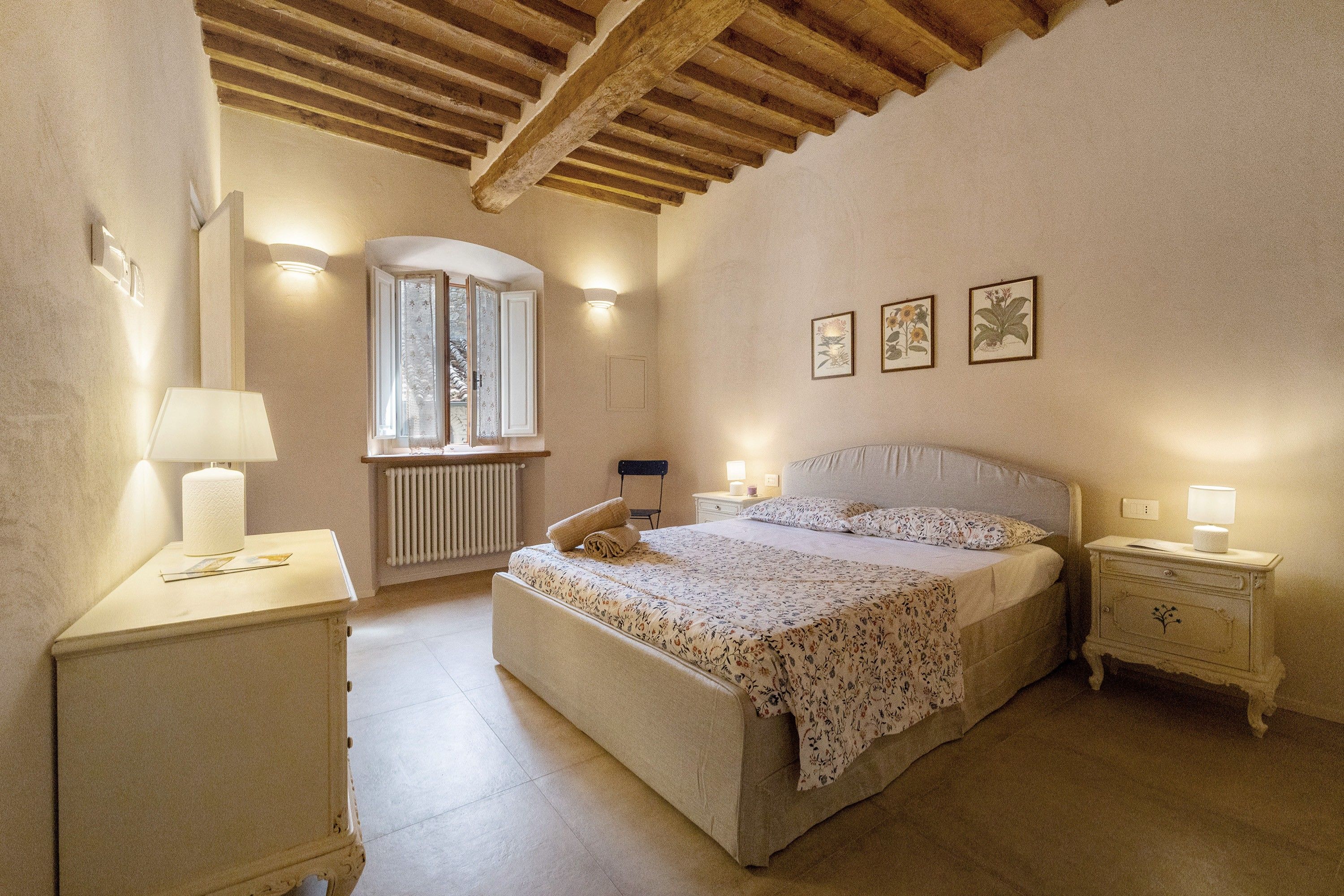 A bedroom with a bed, bedside tables, a radiator, a window with curtains, and wall-mounted lights.