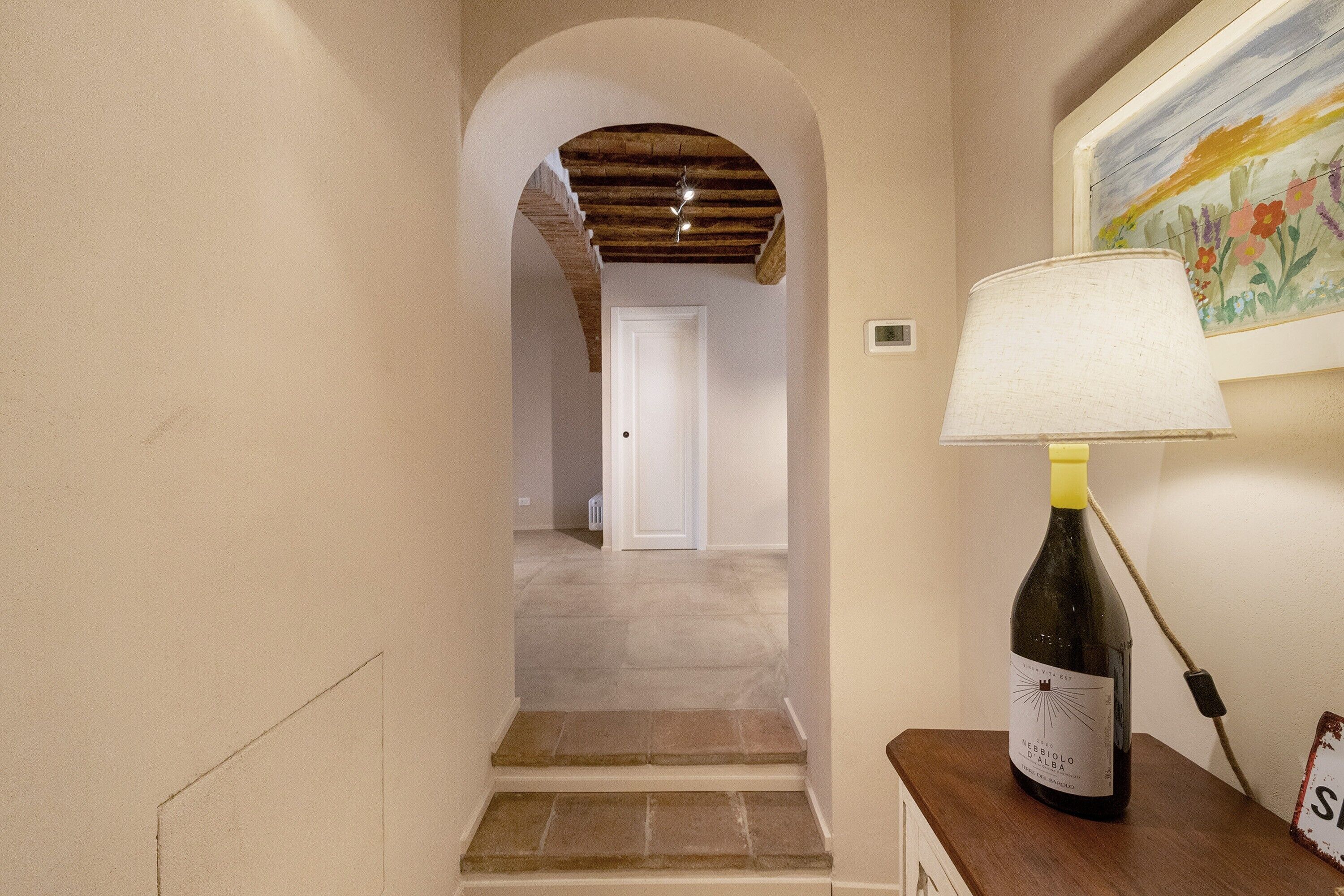 Un couloir avec un escalier menant à une autre pièce, une lampe sur une table et une bouteille de vin.
