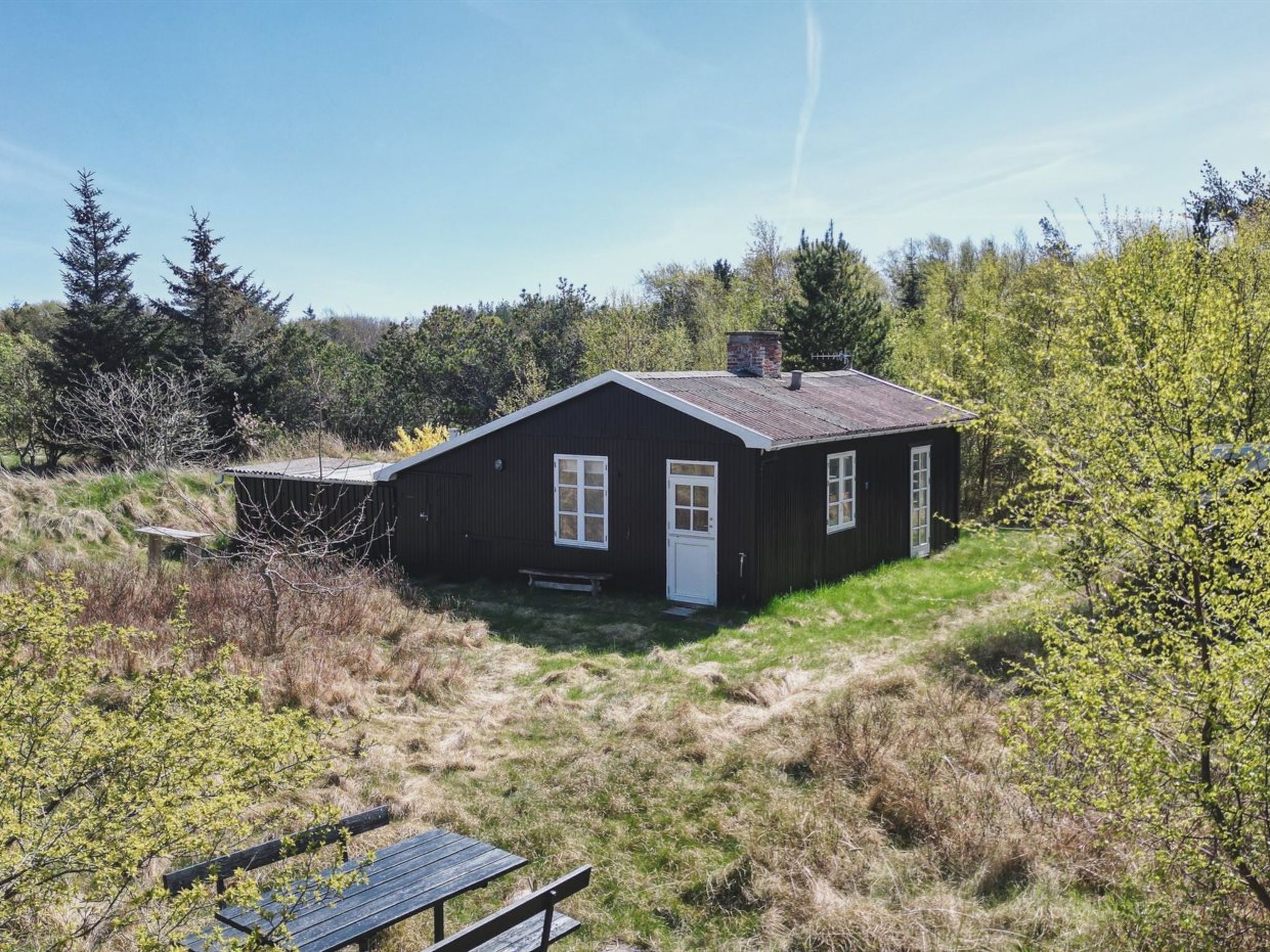 A small, dark wooden house with a white door and windows, surrounded by grass and trees.