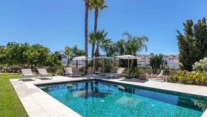 Una piscina rectangular con agua azul cristalina, rodeada por una terraza con baldosas. Hay sillas de descanso y sombrillas, y se ven palmeras al fondo.