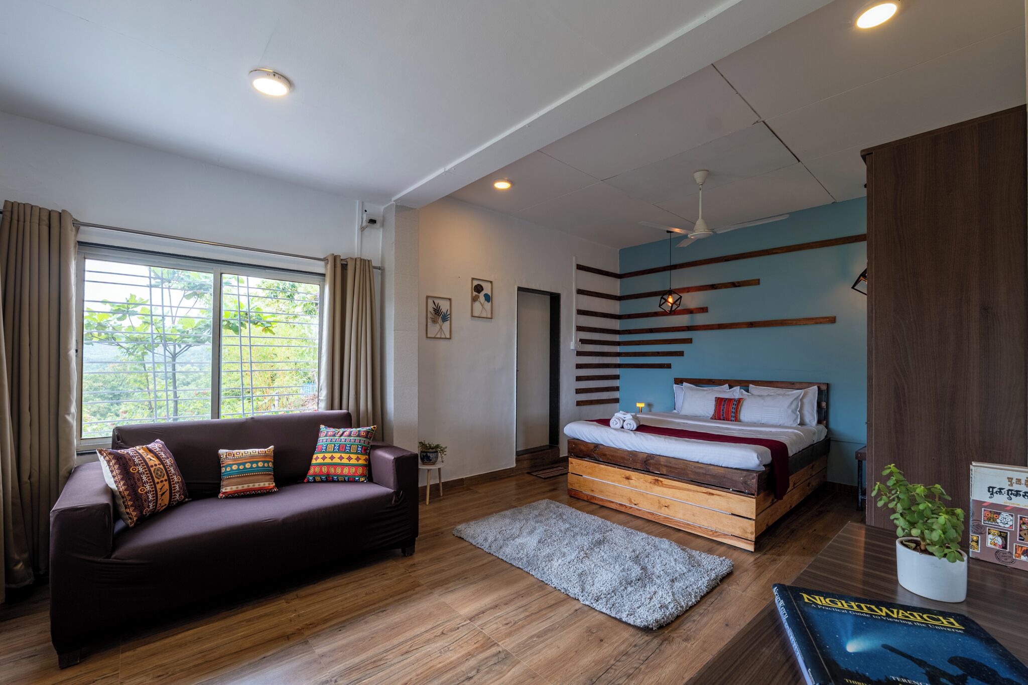 A modern bedroom with a wooden bed, a dark sofa, and a large window.