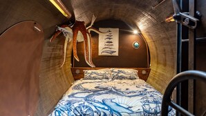 A narrow, tunnel-like room with a bed featuring a nautical-themed bedspread, a wall-mounted poster of marine life, and a copper-colored circular object on the left.