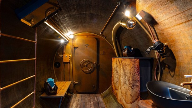 A narrow, metallic-walled room with a wooden floor, a door with a circular emblem, a sink, and a mirror. - Unique lodging, steampunk vibe - 2 guests (Raon-aux-Bois)