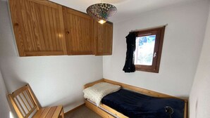 A small, single-bed room with a wooden headboard, a chair, a desk, a window with curtains, and a ceiling light fixture. - Studio at the foot of the slopes - 5 guests (Arêches-Beaufort)