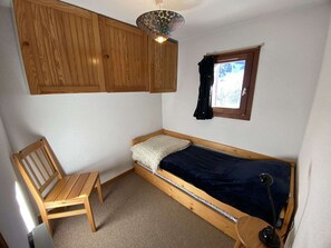 Quarto pequeno com uma cama de solteiro, cabeceira de madeira, uma cadeira, escrivaninha, janela com cortinas e luminária no teto.