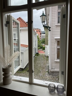 Une vue à travers une fenêtre, avec une porte blanche, un chemin pavé et des bâtiments en arrière-plan.