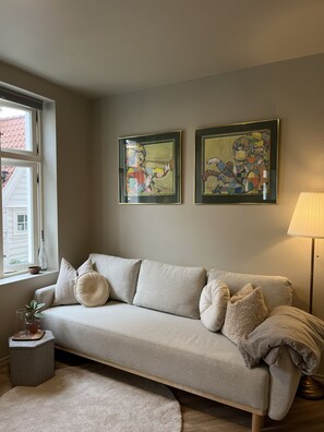 Ett vardagsrum med en neutral färgskala, inrett med en soffa, ett litet sidobord, en lampa och två inramade konstverk på väggen.