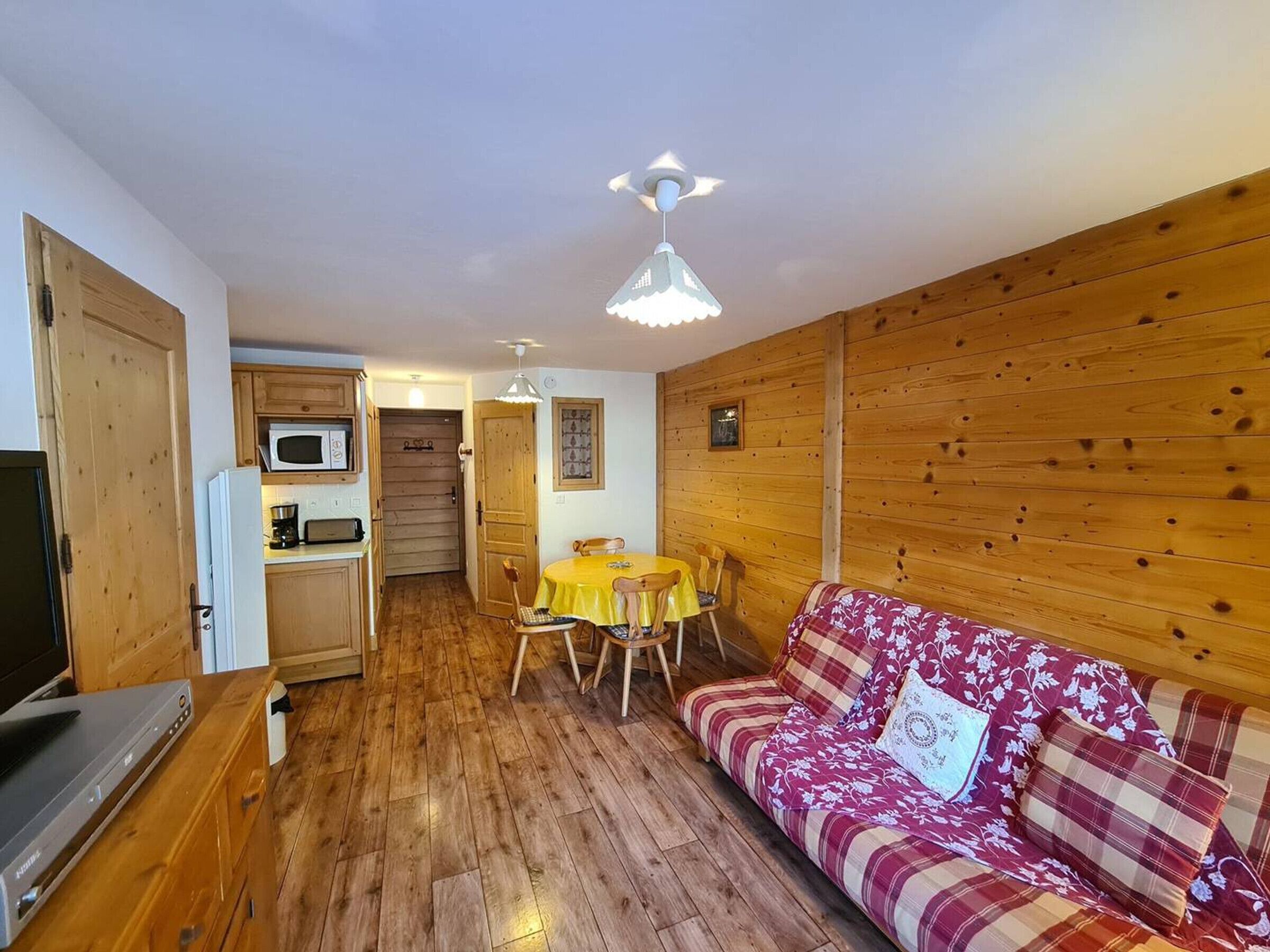 Ein gemütliches Zimmer mit Holzboden, einem Sofa mit floralen und karierten Kissen, einem Esstisch mit Stühlen, einer Mikrowelle und einem Fernseher.
