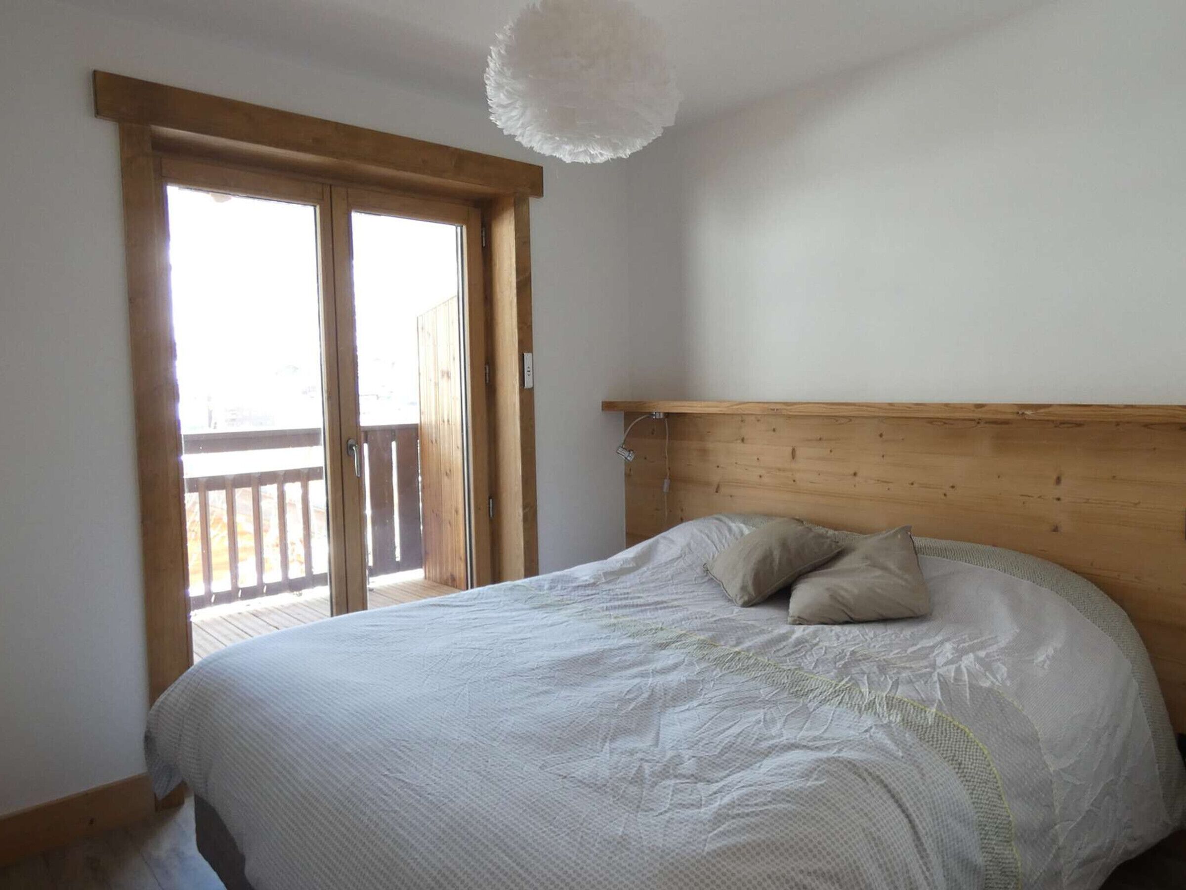 A bedroom with a wooden headboard, a bed with a white duvet and two pillows, a wooden door leading to a balcony, and a white ceiling light.