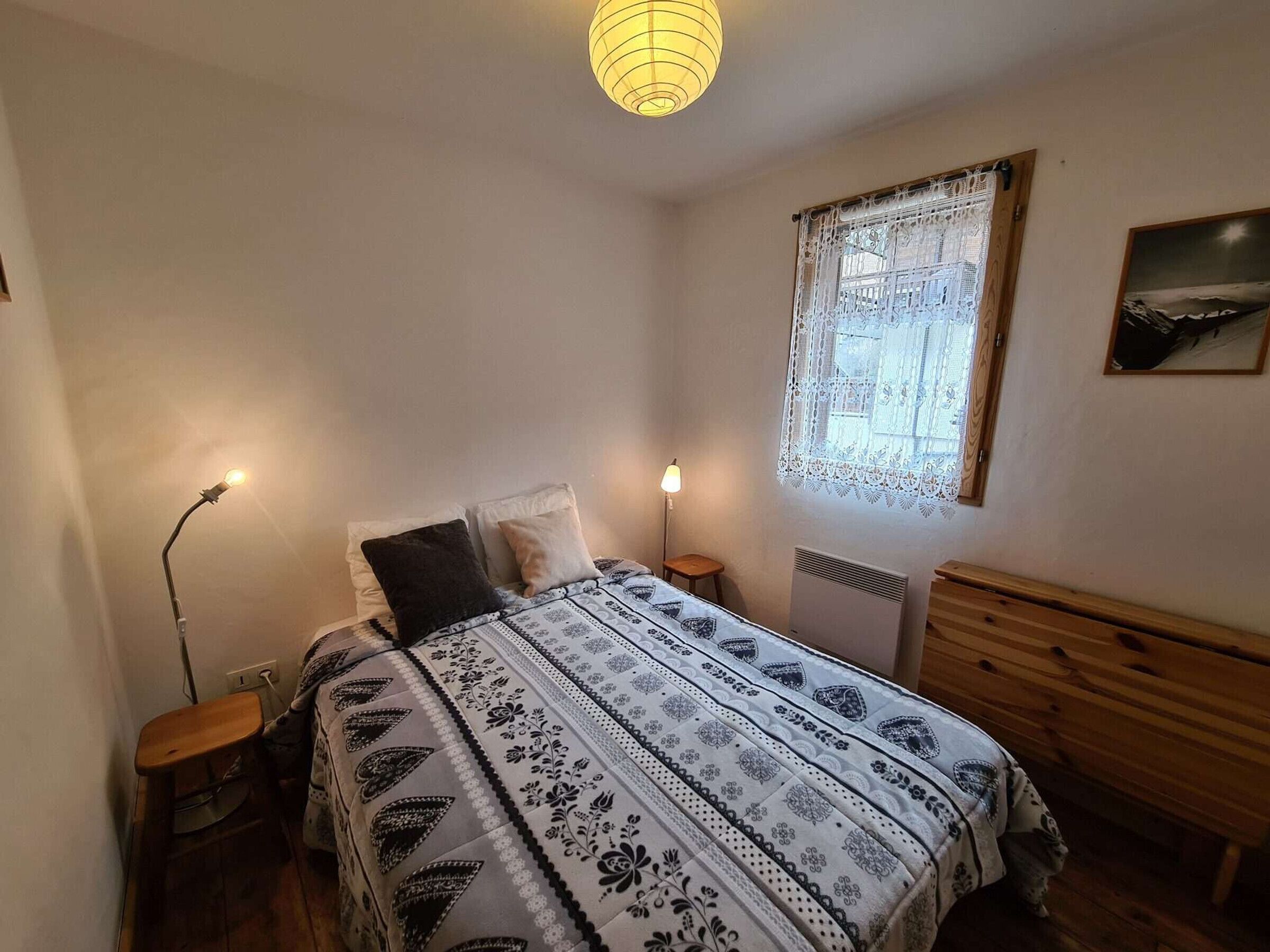 Un dormitorio con cama, lámparas de noche, ventana con cortinas de encaje, radiador y cabecero de madera.