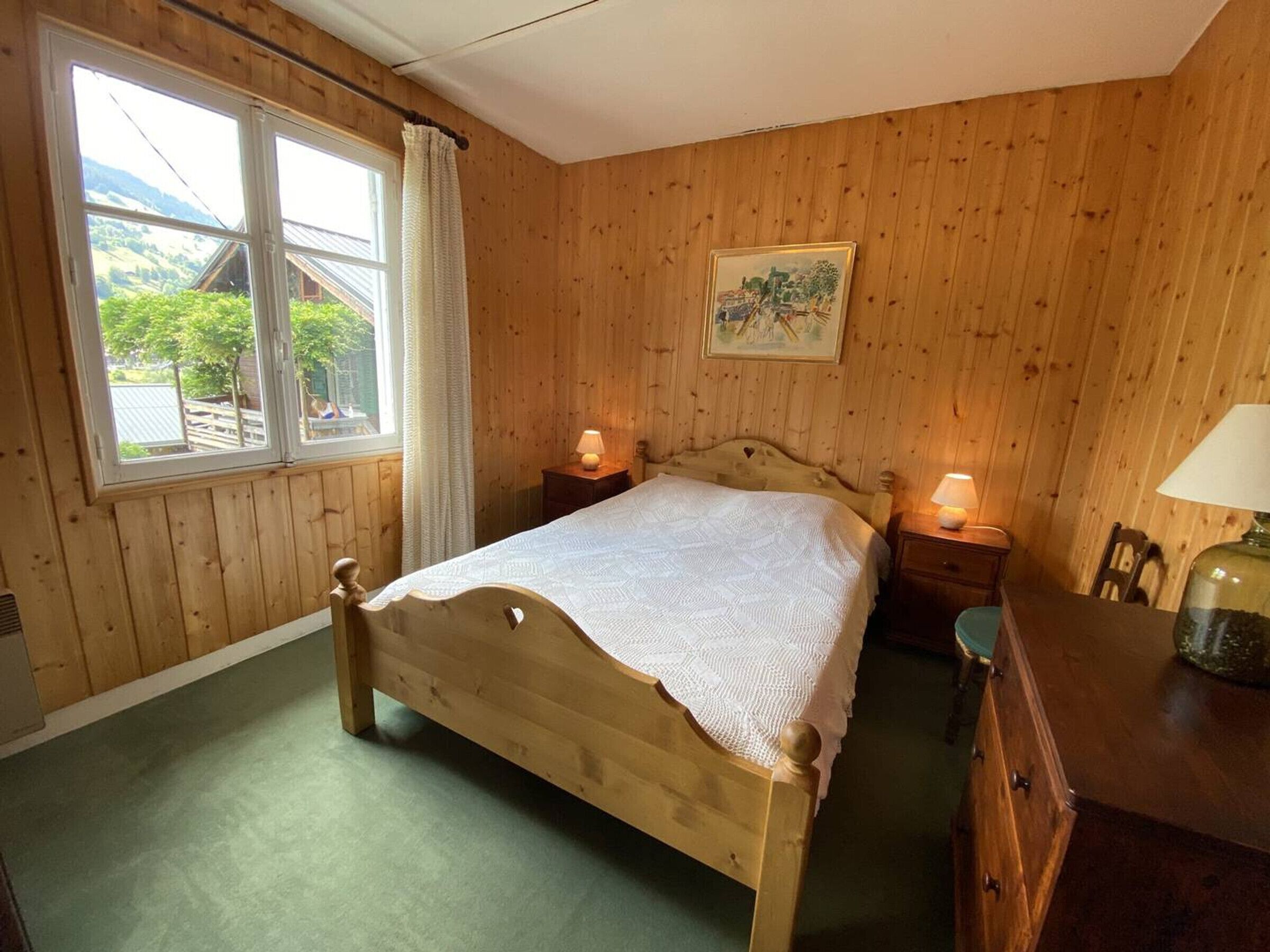 A wooden-paneled bedroom with a bed, bedside lamps, a dresser, and a window with a view of greenery.
