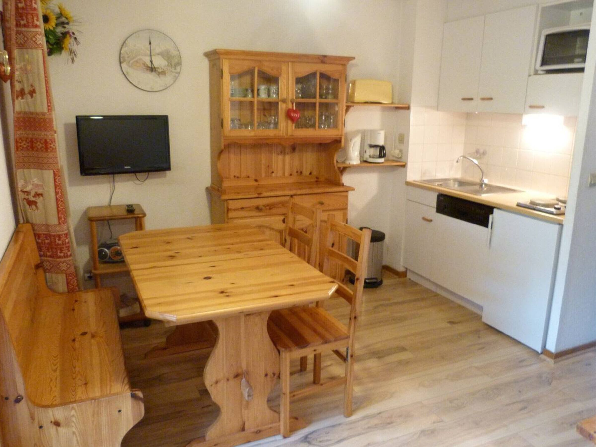 A kitchen with a wooden table, chairs, a cabinet with glass doors, a microwave, and a clock on the wall.