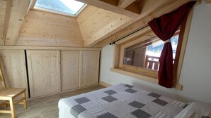 Quarto com teto inclinado, janela com cortina vermelha, cama com colcha e mobiliário de madeira.