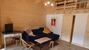 Interior em madeira com sofá, uma mesinha, uma cadeira, televisão em um suporte e luminária no teto.