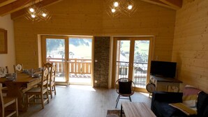 Um quarto espaçoso com paredes de madeira e uma varanda, uma área de jantar com mesa e cadeiras, e uma televisão sobre um suporte.