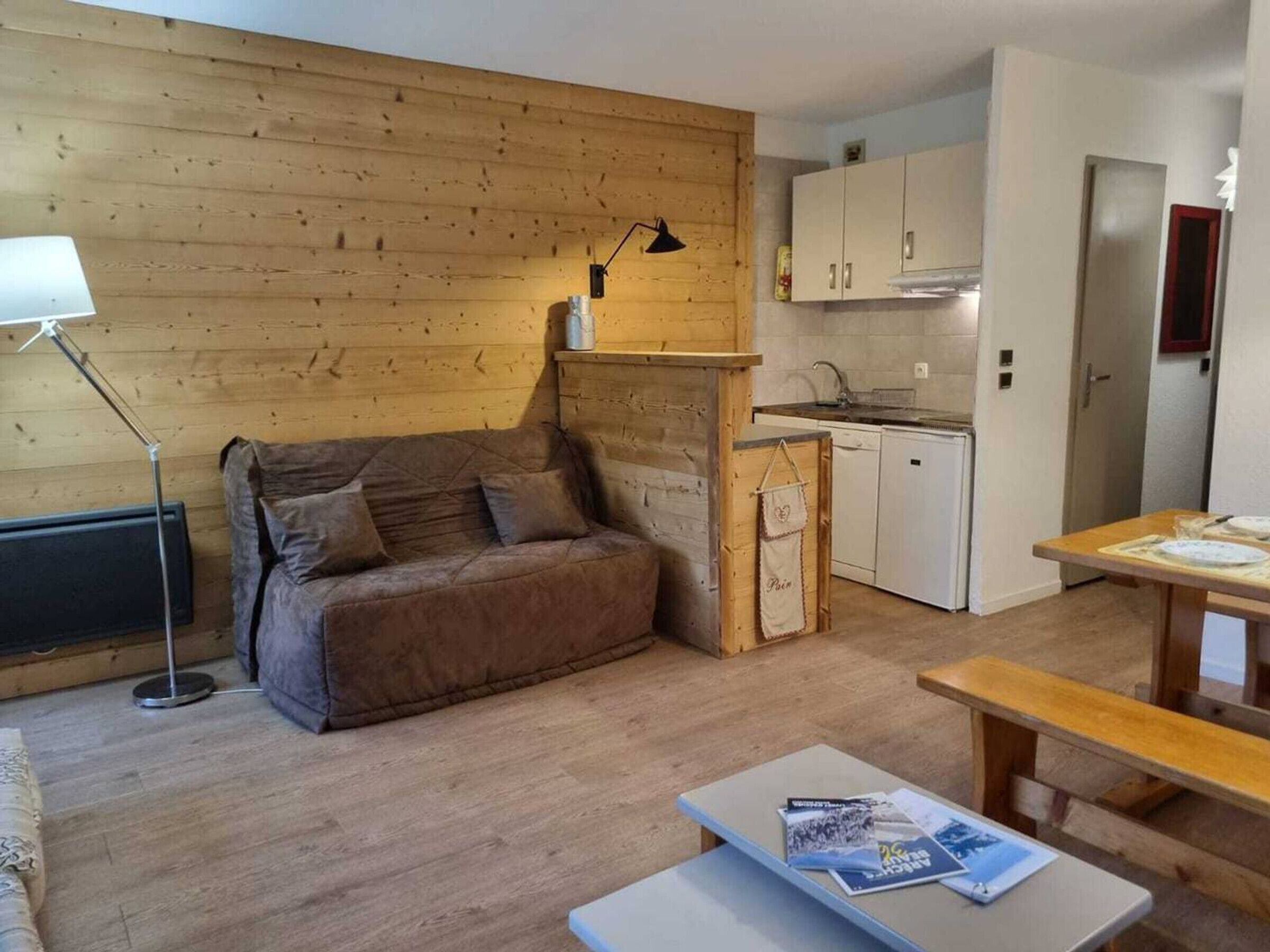 A compact living space with a wooden wall, a sofa, a small table with magazines, a lamp, and a kitchenette.