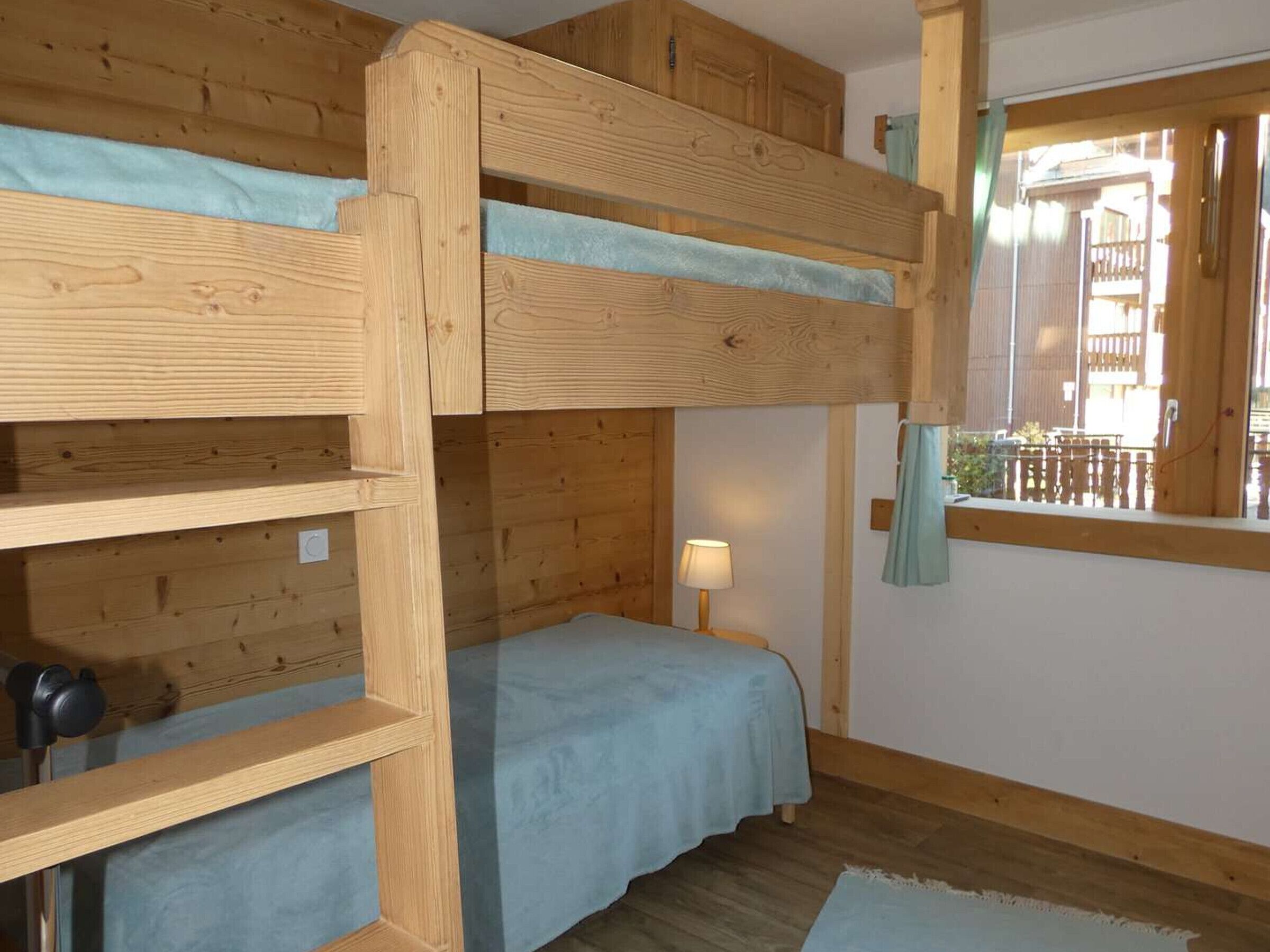 A bunk bed room with wooden walls, a window with a view, and a small bedside table with a lamp.