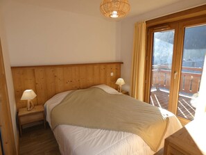 Ein Schlafzimmer mit Holz-Kopfteil, einem Bett mit weißer Bettwäsche, zwei Nachttischlampen und Blick auf eine Holterrasse durch eine Glastür.