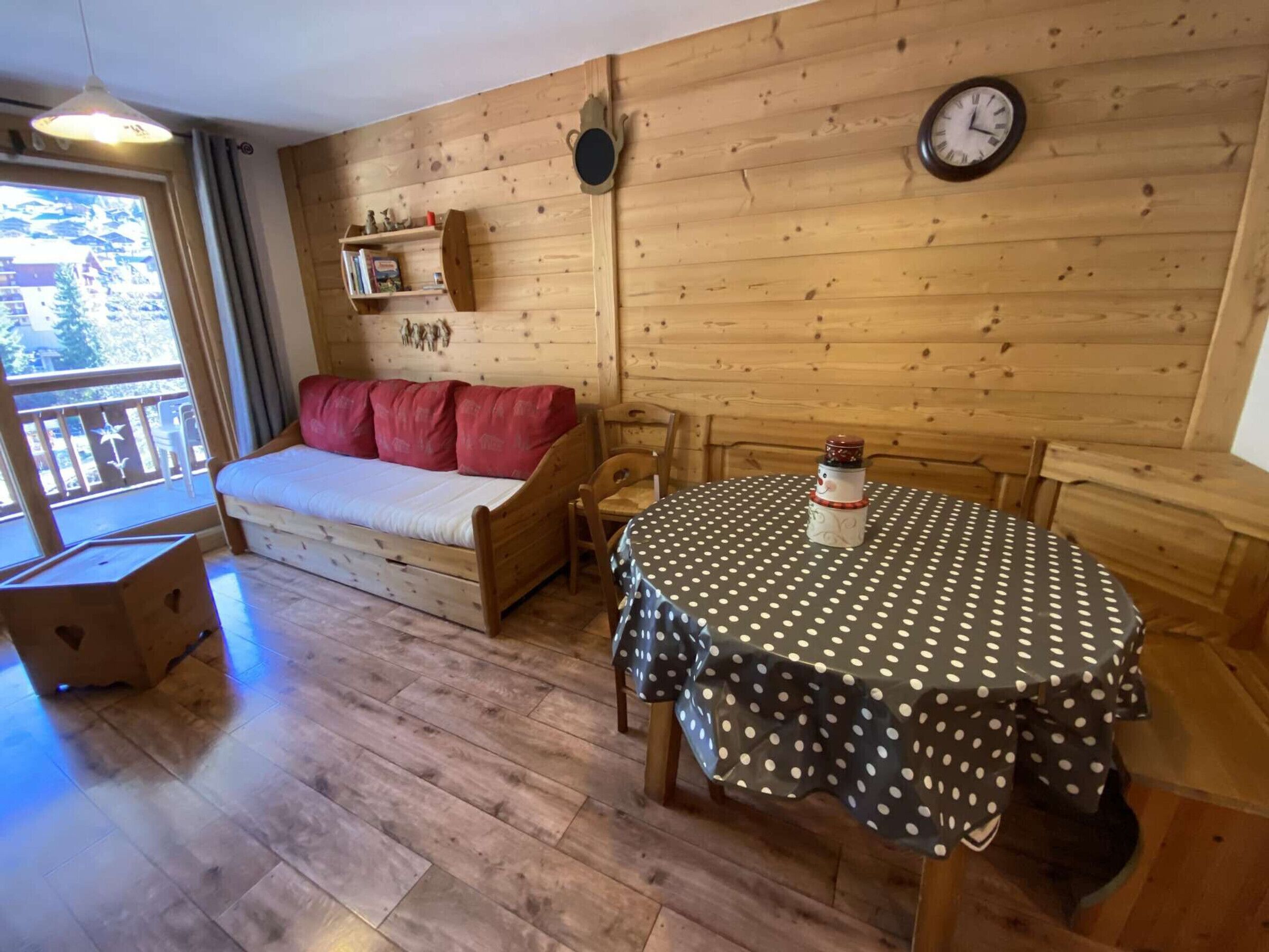 Ein Holzblockhaus-Interieur mit einem Bett, einem Esstisch mit puntiertem Tischlein, einer Uhr an der Wand und einem Fenster mit Blick auf Bäume.